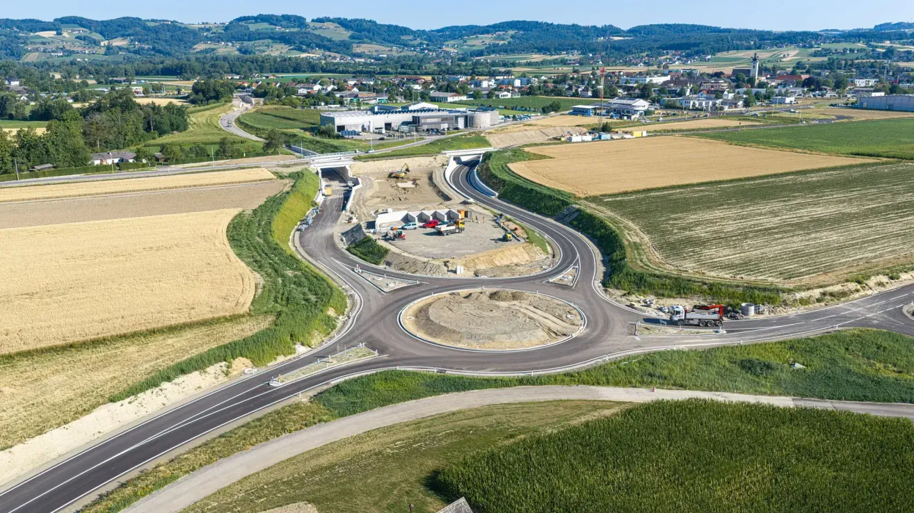 Luftaufnahme einer im Bau befindlichen Kreuzung mit Baumaschinen und Fahrzeugen, umgeben von Feldern und Gebäuden.