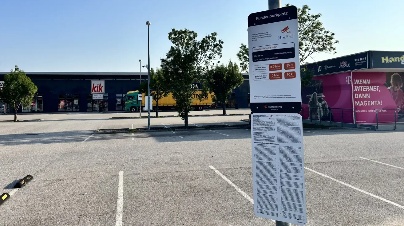 Ein Parkzeichen befindet sich auf einem Parkplatz mit klarem Himmel. Das Schild hat eine digitale Kamera und den Text 'Kundenparkplatz'. Es gibt Bäume und ein Gebäude mit einem Schild, das 'Kik' liest. Vor dem Gebäude ist ein gelber LKW geparkt.