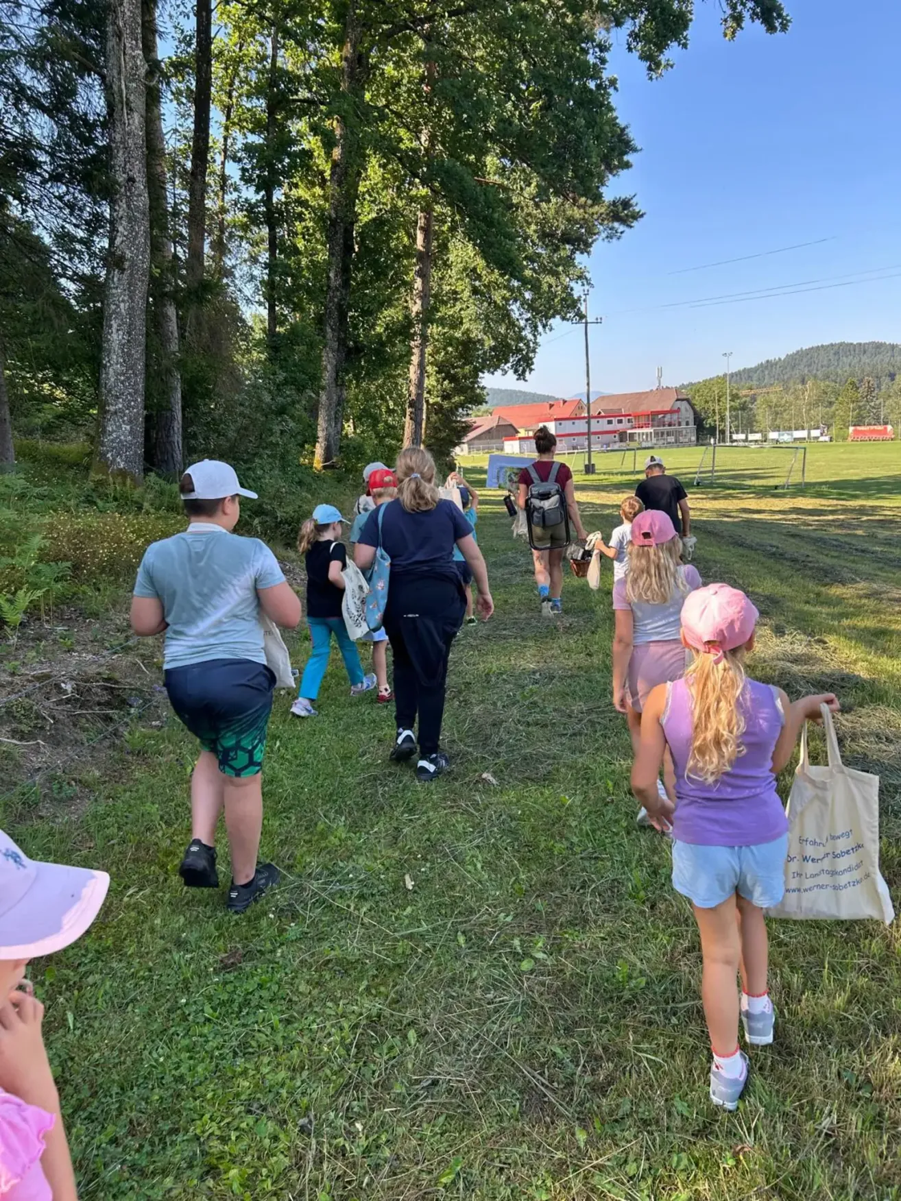 Eine Gruppe von Kindern und Erwachsenen geht auf einem Grasweg in einem Park. Einige tragen Hüte und haben Taschen dabei. Im Hintergrund sind Bäume und ein Gebäude mit rotem Dach zu sehen.