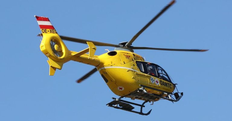 Ein gelber Hubschrauber mit roten und weißen Markierungen fliegt am Himmel und zeigt ein rotes Kreuz und DAMTC an der Seite.