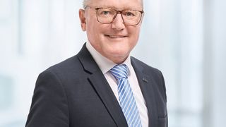Ein Mann in einem Anzug und mit Brille lächelt in die Kamera. Er trägt eine blau-weiß gestreifte Krawatte.