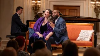 Eine Frau in einem lila Schal singt, während ein Mann in einem blauen Anzug sie festhält, auf einer orangefarbenen Couch vor einem Klavier sitzend.