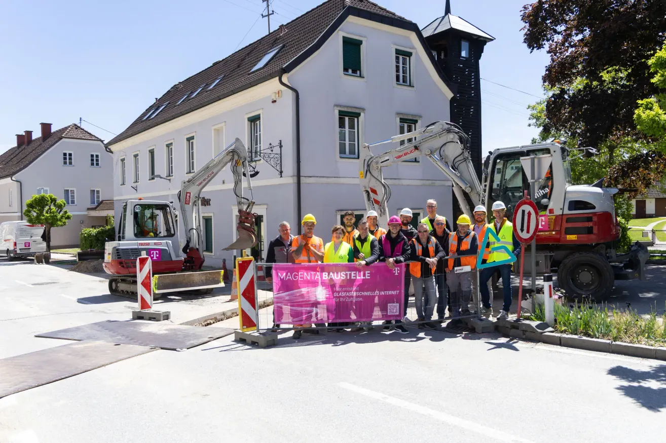 Eine Gruppe von Arbeitern in Warnwesten und Helmen steht hinter einem Banner mit der Aufschrift Magenta Baustelle, mit Baugeräten und einem großen Gebäude im Hintergrund.