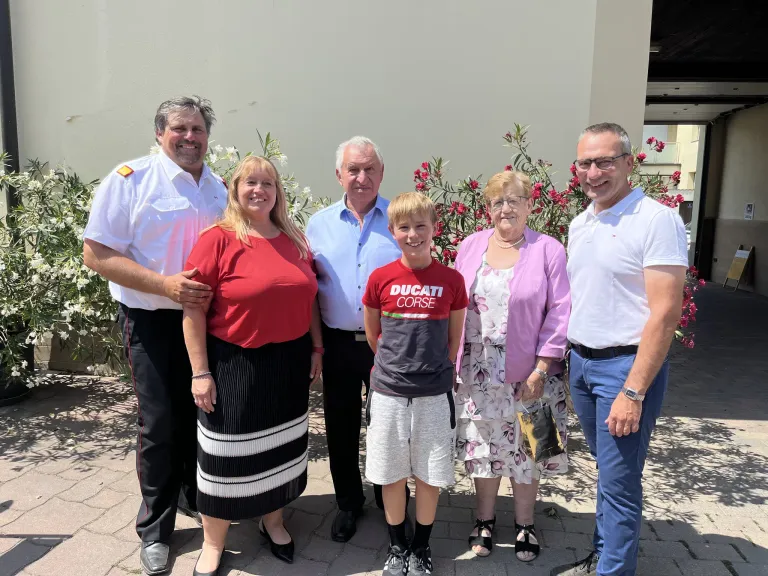 Ein Familienfoto mit sechs Personen vor einer weißen Wand mit roten Blumen. Der Mann links trägt eine Uniform und die Frau neben ihm ein rotes Shirt. Der Junge in der Mitte trägt ein rotes Shirt mit dem Wort Ducati.