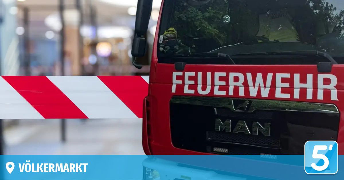 Ein roter Feuerwehrwagen mit dem Wort 'FEUERWEHR' auf der Front, geparkt vor einem verschwommenen Hintergrund mit einem rot-weiß gestreiften Hindernis.