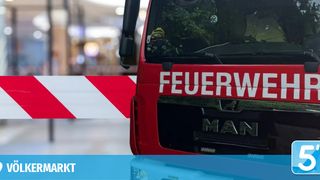 Ein roter Feuerwehrwagen mit dem Wort 'FEUERWEHR' auf der Front, geparkt vor einem verschwommenen Hintergrund mit einem rot-weiß gestreiften Hindernis.