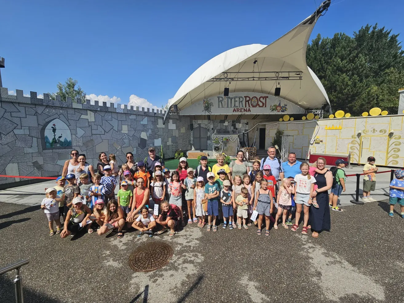 Eine Gruppe von Kindern und Erwachsenen posiert für ein Foto vor einer burgartigen Bühne mit weißem Vordach. Die Bühne hat ein Schild mit der Aufschrift Ritterrost Arena.