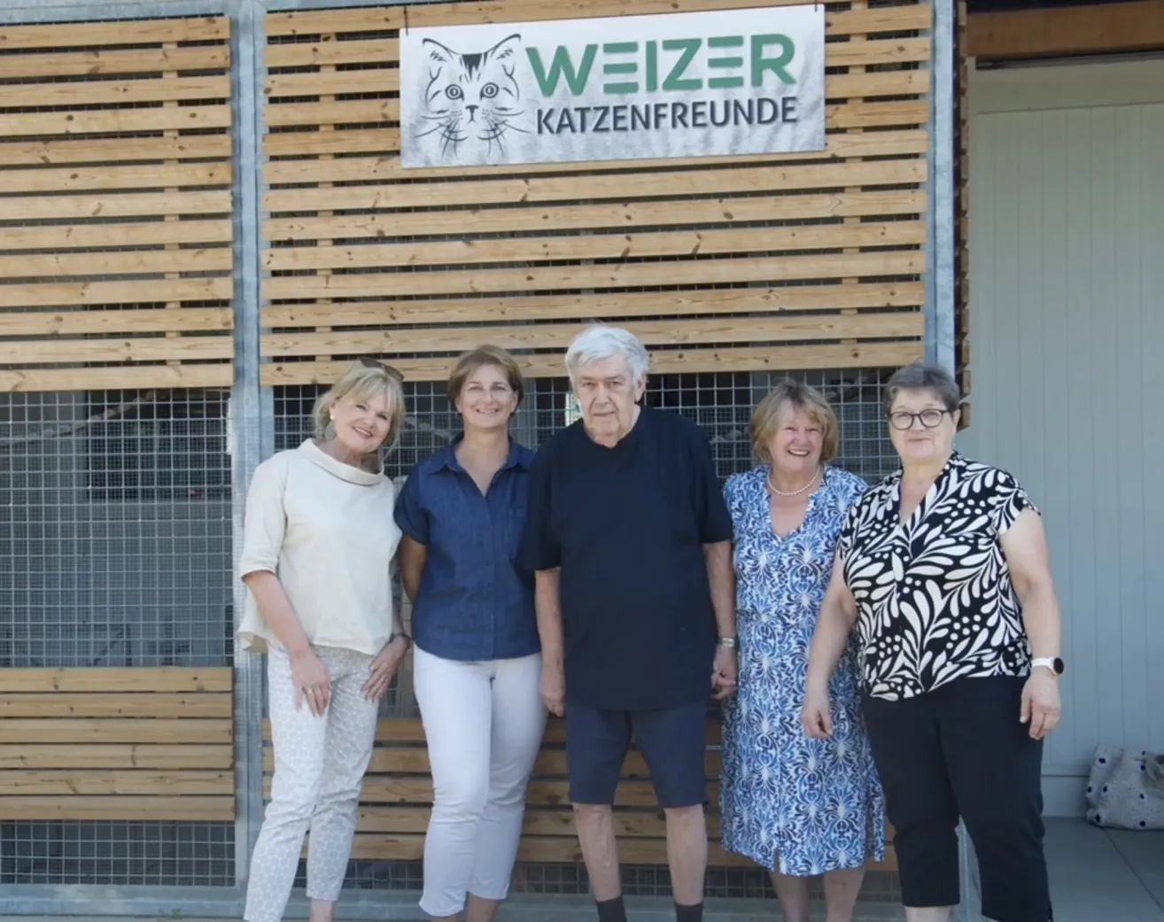 Fünf Personen stehen vor einer Holzwand mit einem Schild, das 'Weizer Katzenfreunde' besagt. Die Gruppe umfasst einen Mann mit weißen Haaren und eine Frau mit Brille.