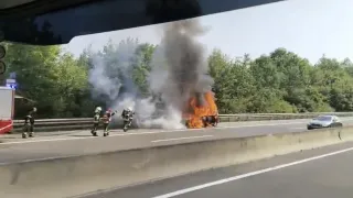 Feuerwehrleute löschen ein brennendes Fahrzeug auf einer Autobahn mit Bäumen im Hintergrund.