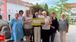 Eine Gruppe von Menschen steht zusammen, lächelt und hält einen gelben Scheck. Die Frau in der Mitte hält den Scheck mit beiden Händen, und die anderen stehen hinter ihr. Sie sind alle lässig gekleidet.