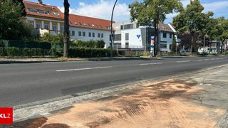 Eine leere Straße mit einem Bürgersteig und einem Gebäude mit rotem Dach und weißen Wänden. Bäume und Büsche stehen vor dem Gebäude. Ein Pfosten mit einem Schild steht auf dem Bürgersteig. Ein Auto ist am Straßenrand geparkt.
