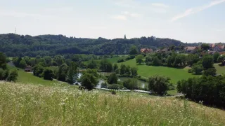 Eine friedliche ländliche Landschaft mit einem Fluss, der sich durch saftige grüne Felder schlängelt, und Hügeln im Hintergrund.