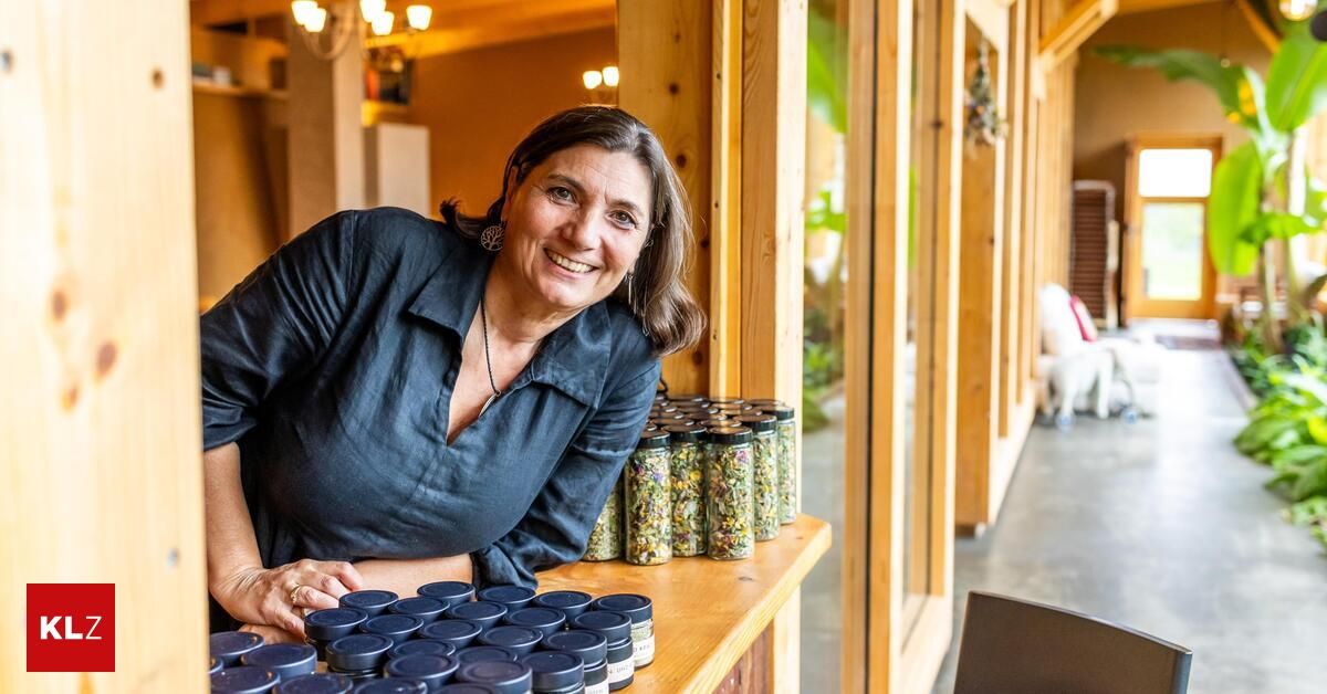 Eine Frau lächelt und lehnt sich an einen Holztresen, der mit Gläsern voller Kräuter gefüllt ist.