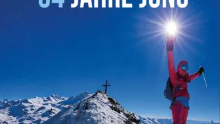 Eine Person steht auf einem schneebedeckten Berggipfel, hält einen Skistock und eine Lampe und die Worte '54 Jahre alt, 34 Jahre jung' darüber.