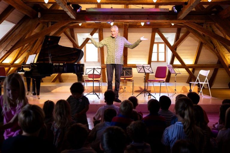 Ein Mann steht auf einer Bühne mit einem Klavier und Notenständern und spricht eine Gruppe von Kindern an. Die Bühne ist mit warmer Beleuchtung und Holzbalken ausgestattet.