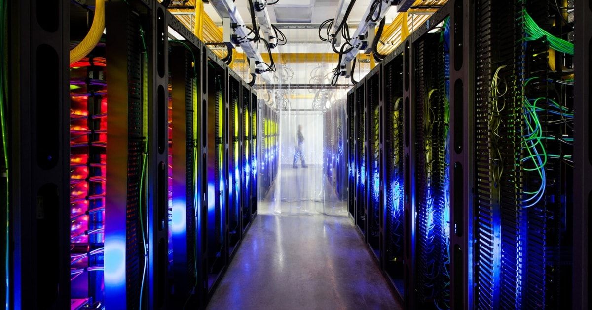 Ein Serverraum gefüllt mit Reihen von Server-Racks, beleuchtet mit blauen und gelben Lichtern, mit einer Person in der Ferne stehend.