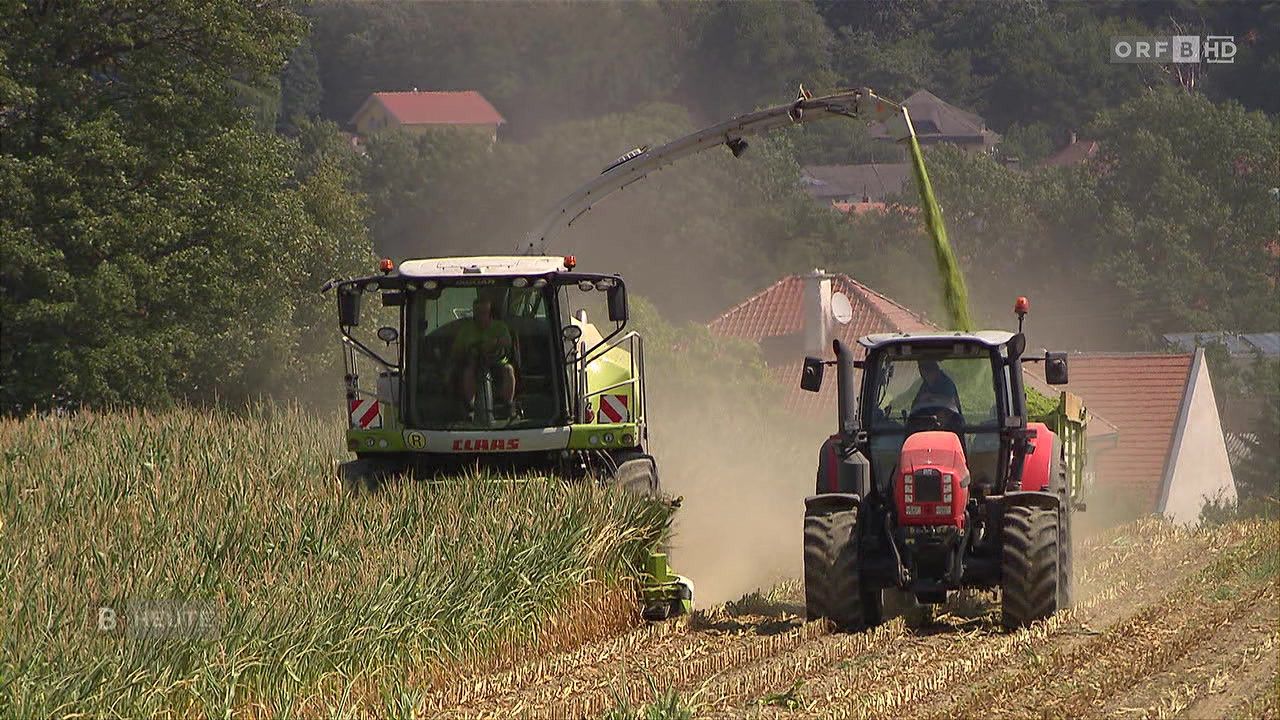 Ein Traktor mit einem großen Anbaugerät erntet Feldfrüchte, während ein weiterer Traktor dicht dahinter folgt. Die Szene spielt in einer ländlichen Gegend mit Häusern im Hintergrund.