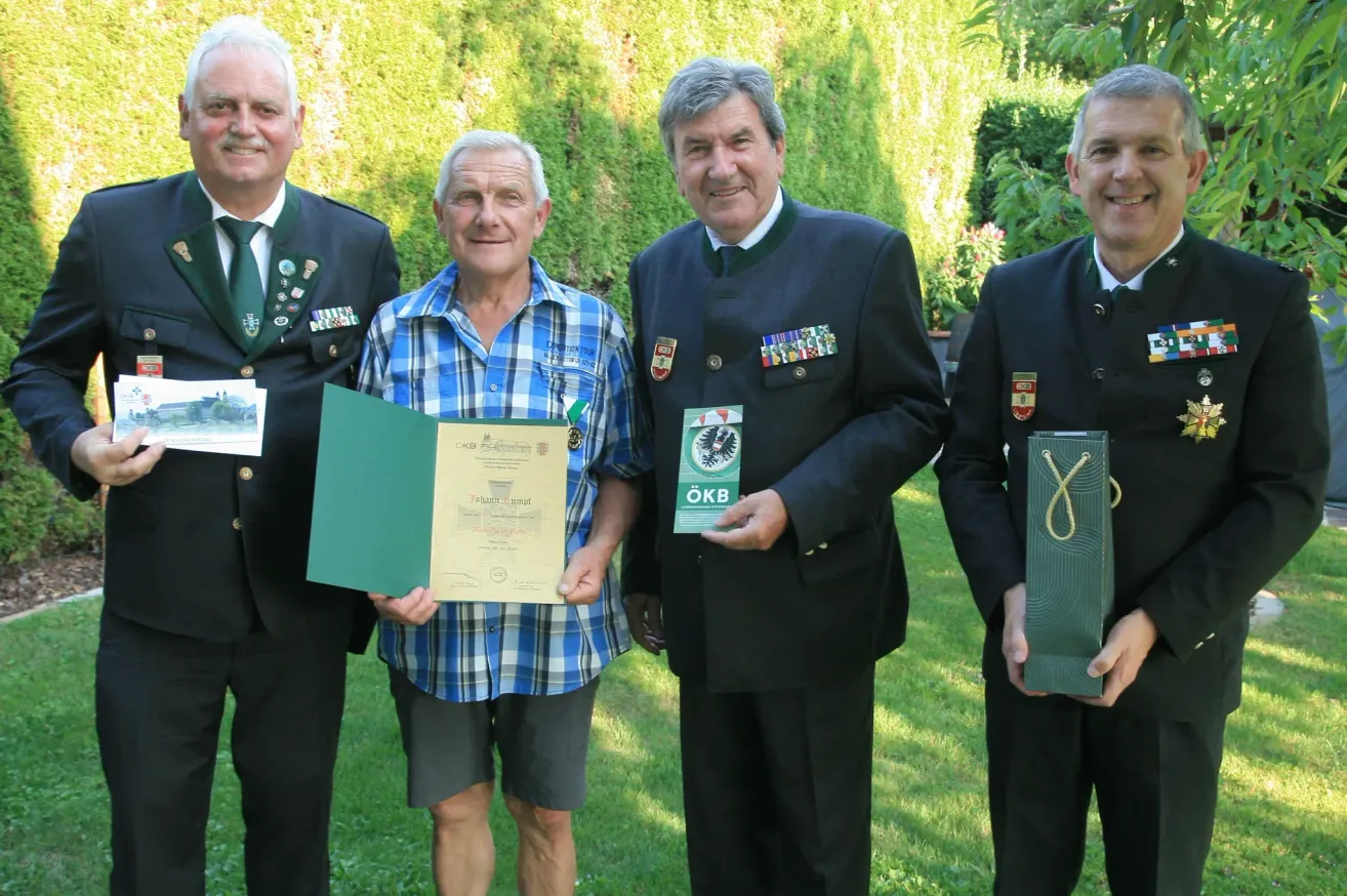 Vier Männer stehen auf einem Rasen, lächelnd, einer hält ein Zertifikat und ein anderer eine grüne Tasche. Sie tragen Uniformen und Medaillen.