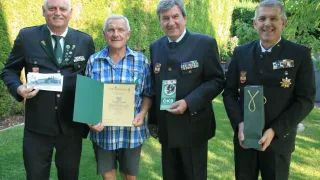 Vier Männer stehen auf einem Rasen, lächelnd, einer hält ein Zertifikat und ein anderer eine grüne Tasche. Sie tragen Uniformen und Medaillen.