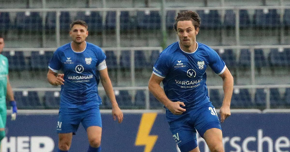 Two soccer players in blue uniforms are running on the field. One player is looking to the left, and the other is looking forward. Behind them is a blurry background of empty blue chairs.