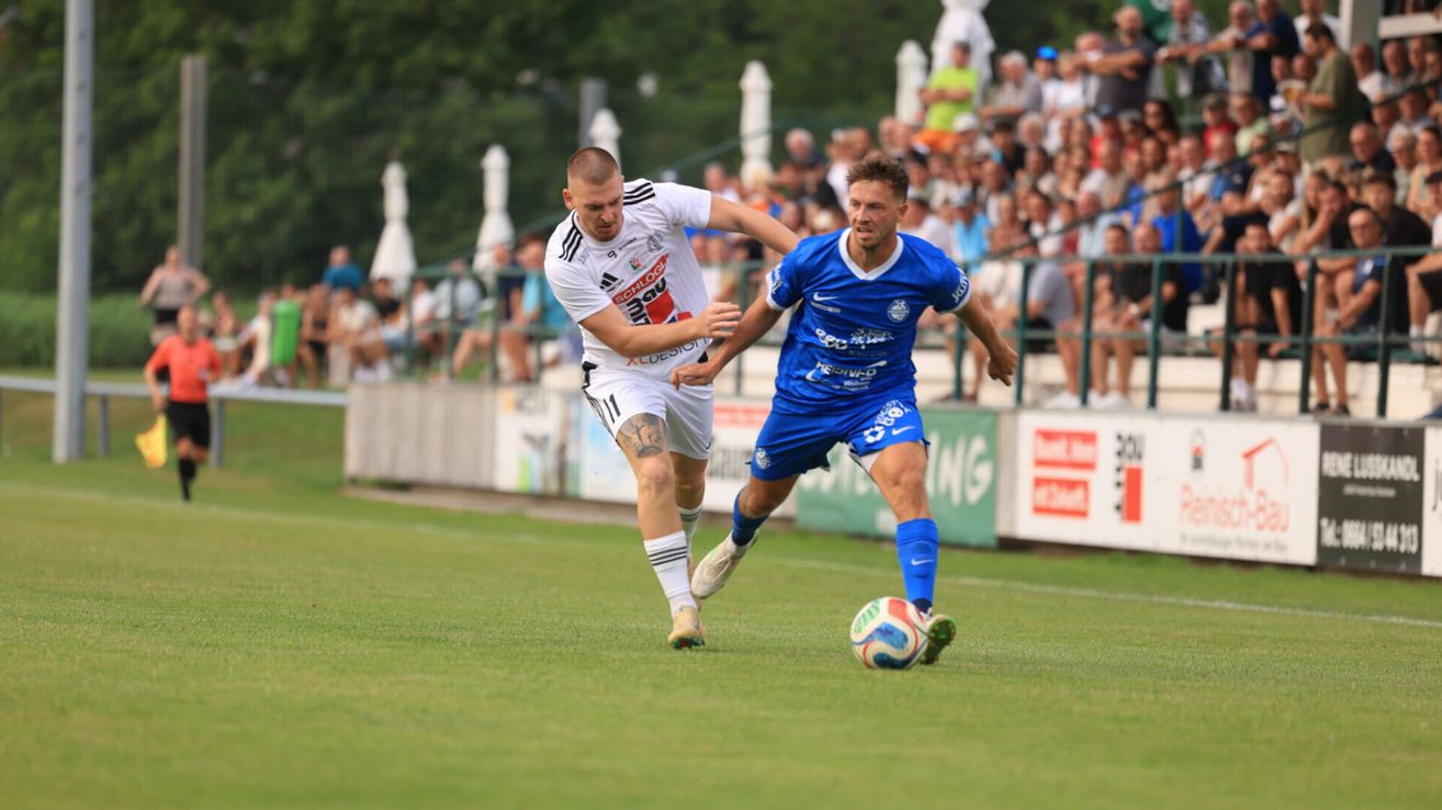 Zwei Fußballspieler, einer in Weiß und der andere in Blau, kämpfen auf einem Feld um den Ball. Zuschauer schauen von den Tribünen zu.