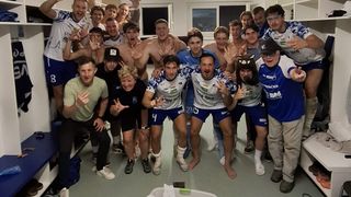 Eine Gruppe von Athleten in einer Umkleidekabine, einige in Sportuniformen, posiert für ein Foto. Sie alle lächeln und machen Handgesten.
