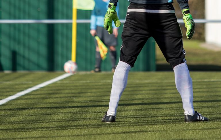 Ein Fußballspieler in schwarz-weißem Trikot mit grünen Handschuhen steht auf dem Feld. Eine verschwommene Figur eines Schiedsrichters hält eine gelbe Flagge.