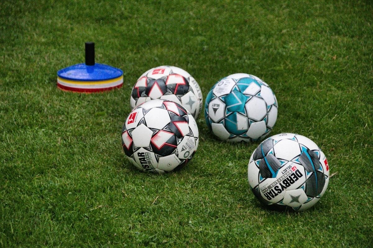 Five soccer balls of different designs are placed on a green grass field with a blue and red cone in the background.