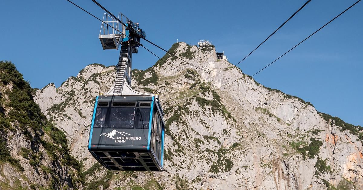 A cable car labeled 'Untersberg Bahn' is ascending a steep mountain with a clear blue sky in the background.