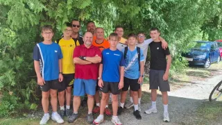 Eine Gruppe von Männern und Jungen in Sportkleidung posiert für ein Foto unter einem Baum. Einige tragen eine Brille und Turnschuhe, und ein Mann hat die Arme verschränkt.