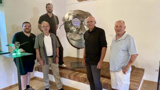 Fünf Männer stehen vor einer metallischen Skulptur und lächeln für ein Foto. Die Skulptur ist groß und metallisch, mit einer gesichtsähnlichen Struktur. Die Männer sind lässig gekleidet.