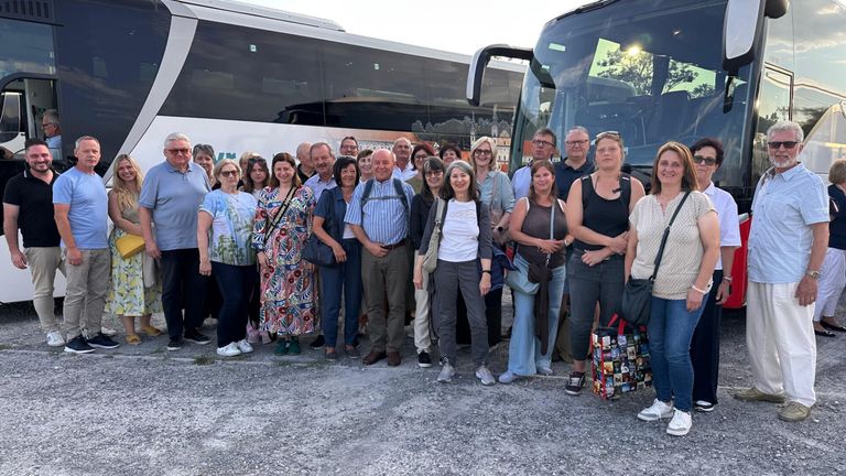 Eine Gruppe von Menschen steht vor zwei Bussen und lächelt für ein Foto. Sie sind lässig gekleidet und einige tragen Taschen. Die Umgebung scheint im Freien zu sein.