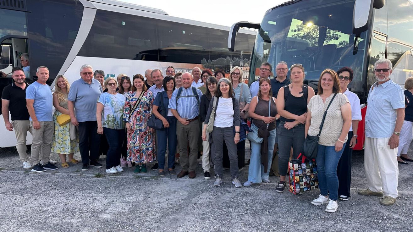 Eine Gruppe von Menschen steht vor zwei Bussen und lächelt für ein Foto. Sie sind lässig gekleidet und einige tragen Taschen. Die Umgebung scheint im Freien zu sein.