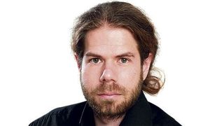 Ein Mann mit Bart und Schnurrbart in einem schwarzen Hemd, der direkt in die Kamera mit einem neutralen Ausdruck schaut.