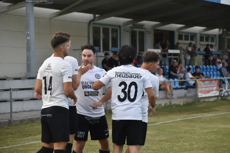 Eine Gruppe von Fußballspielern in weißen Trikots steht auf einem Feld und diskutiert wahrscheinlich über das Spiel. Der Spieler in der Mitte trägt ein Trikot mit der Nummer 30. Hinter ihnen sitzen Menschen auf Tribünen.
