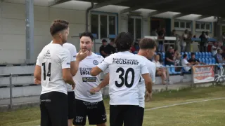 Eine Gruppe von Fußballspielern in weißen Trikots steht auf einem Feld und diskutiert wahrscheinlich über das Spiel. Der Spieler in der Mitte trägt ein Trikot mit der Nummer 30. Hinter ihnen sitzen Menschen auf Tribünen.