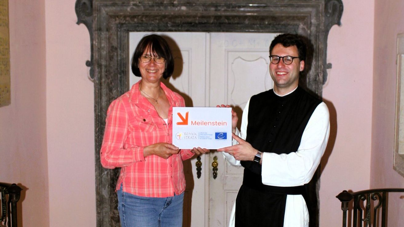 A woman in a pink shirt and a man in a black vest hold a certificate with the name Meilenstein on it. They are standing in front of a white door.