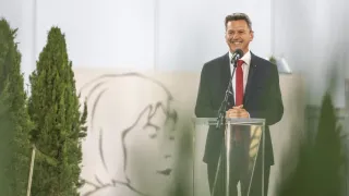 Ein Mann in Anzug und Krawatte steht lächelnd an einem Rednerpult, mit einem Mikrofon und einer Zeichnung einer Frau hinter ihm.
