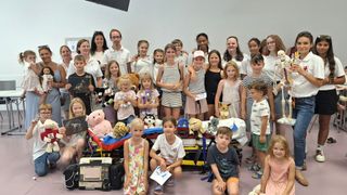 Eine Gruppe von Kindern und Erwachsenen posiert für ein Foto mit Kuscheltieren und einer Trage in einem Raum.
