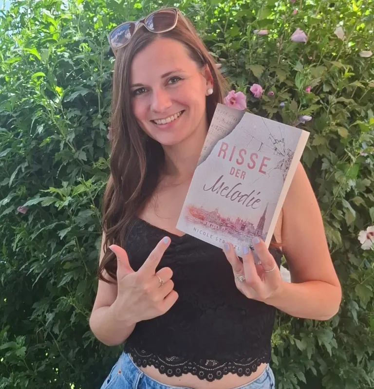 Eine Frau mit Sonnenbrille auf dem Kopf und einem Ring am Finger lächelt und hält ein Buch mit dem Titel 'Risse der Melodie' vor einem grünen Busch.