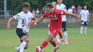 Zwei Fußballspieler in Aktion auf einem Feld, einer in Rot und der andere in Weiß, mit einem verschwommenen Hintergrund von Zuschauern und Bäumen.