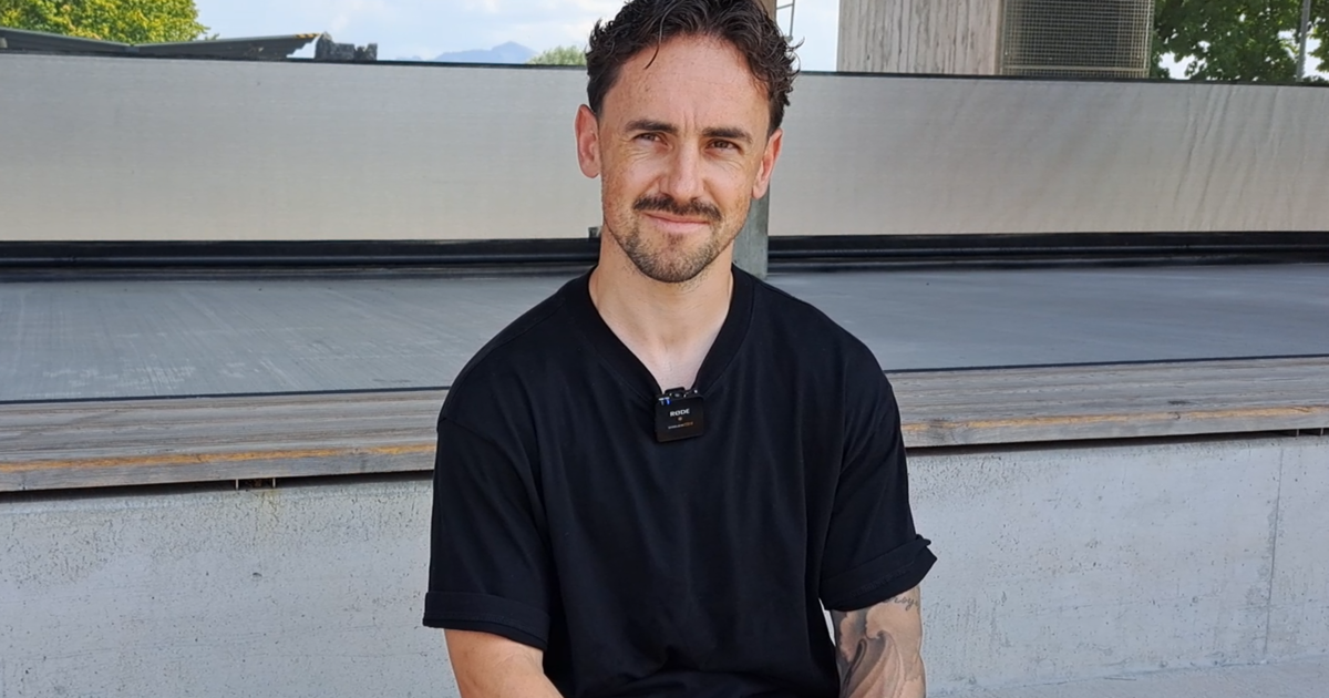 A man with a beard and mustache wearing a black shirt with a microphone attached to his collar, sitting on a concrete bench.