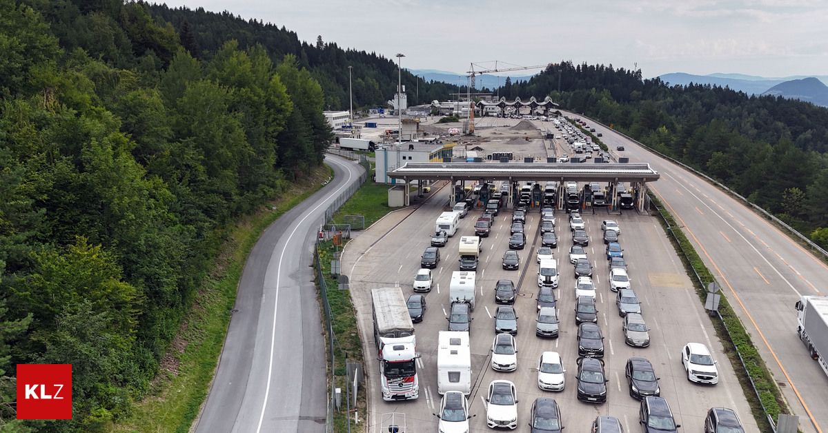 Luftaufnahme einer geschäftigen Mautstelle mit zahlreichen Fahrzeugen, darunter Autos, Lastwagen und Lieferwagen, die in mehreren Spuren warten, um zu bezahlen. Die Mautstelle ist von viel Grün umgeben, und in der Ferne sind Berge zu sehen.
