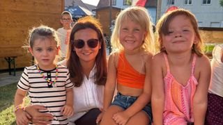 Eine Frau mit langen braunen Haaren und Sonnenbrille lächelt mit drei jungen Mädchen, eines mit einer bunten Halskette, ein anderes in einem orangefarbenen Tanktop und das dritte in einem rosa Kleid.