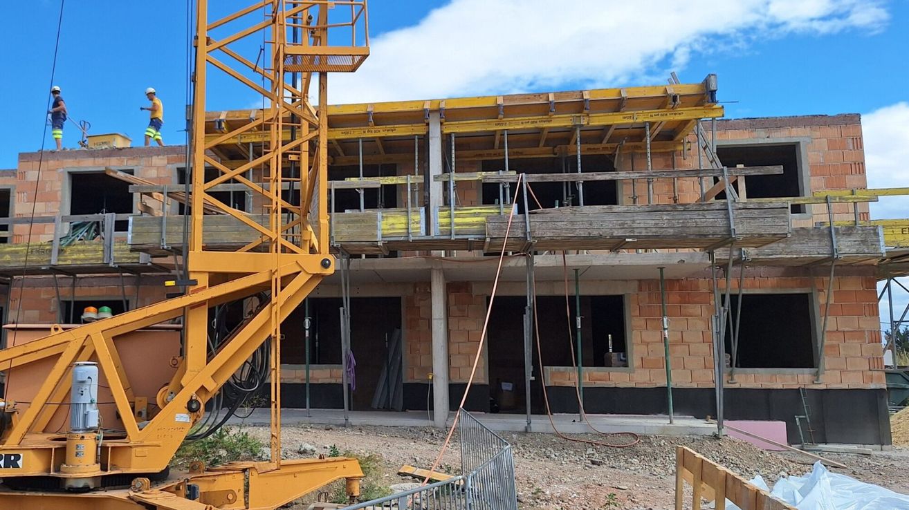 Eine Baustelle mit einem gelben Kran und Gerüsten, die ein unfertiges Gebäude mit Ziegelwänden und offenen Fenstern zeigt.