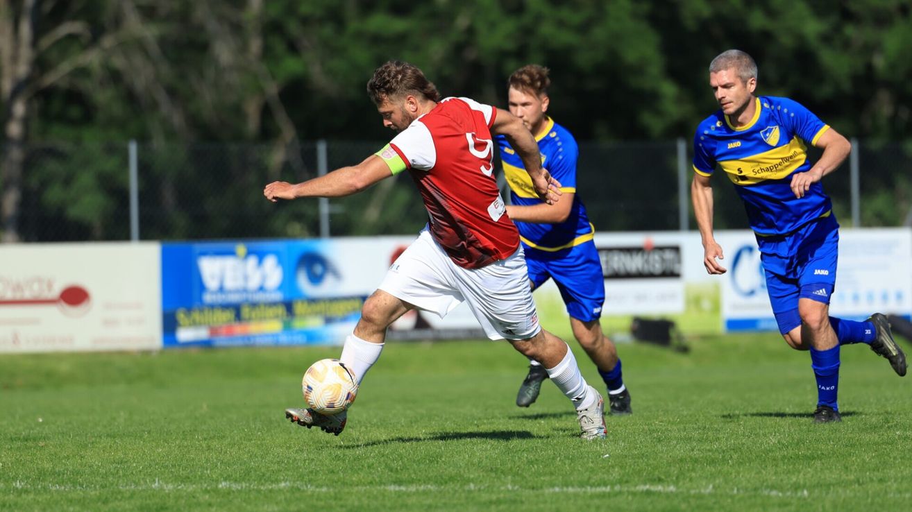 Ein Fußballspieler in einem roten Trikot mit der Nummer 3 tritt einen Ball auf einem Feld, hinter ihm ein weiterer Spieler in Blau.
