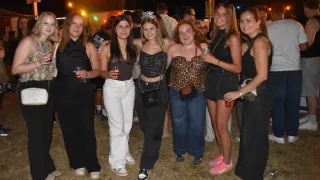 Eine Gruppe von Frauen in verschiedenen Outfits steht bei einem nächtlichen Outdoor-Event zusammen, lächelt und hält Getränke. Eine Frau trägt eine Geburtstagskrone.