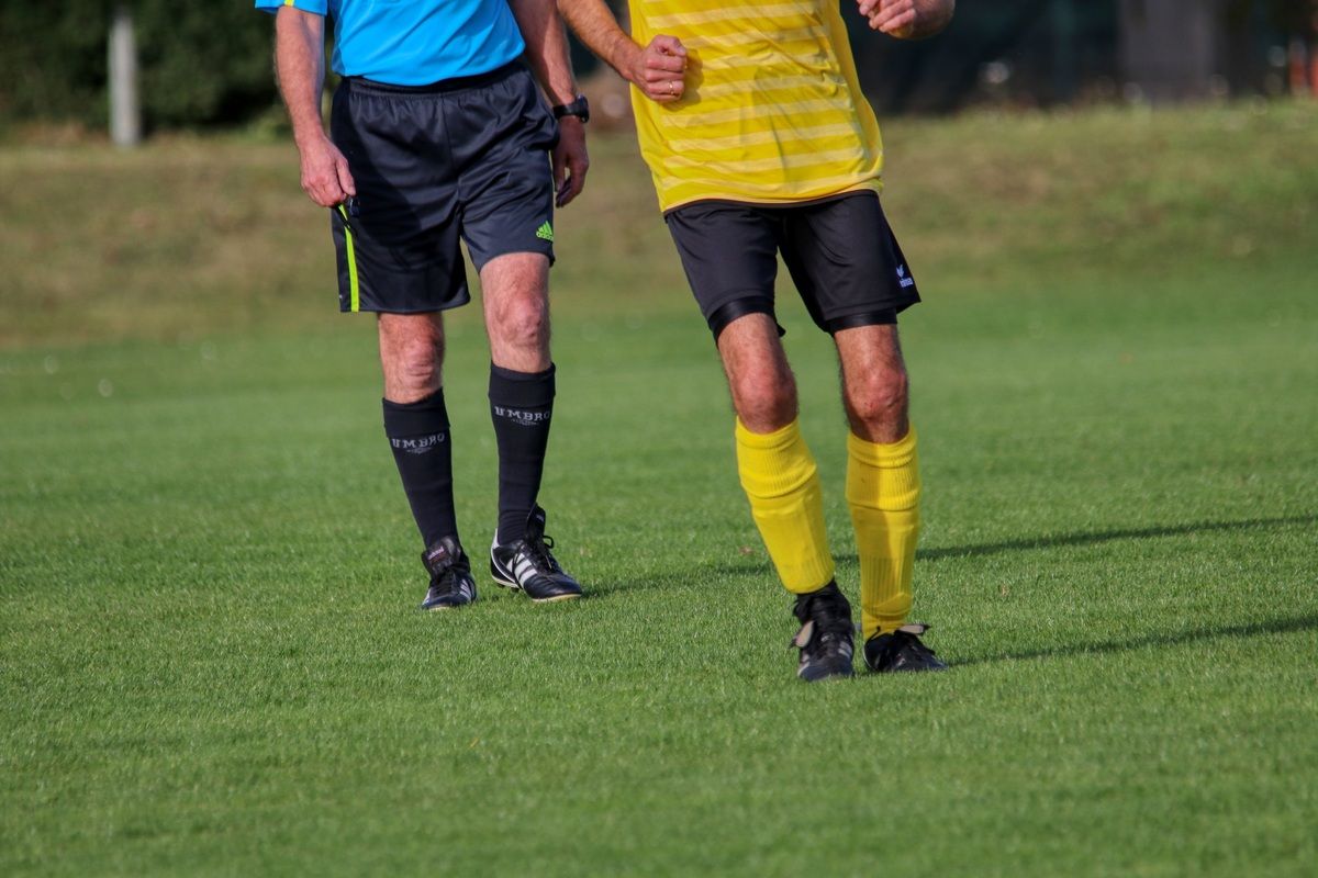 Zwei Männer auf einem Fußballfeld, einer in einem blauen Shirt und schwarzen Shorts, der andere in einem gelben Shirt und schwarzen Shorts.