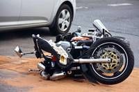 Ein Motorrad liegt auf der Seite auf der Straße, mit abgekoppeltem Vorderrad. Ein silbernes Auto ist in der Nähe geparkt, und die Straße besteht aus Asphalt.