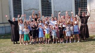 Eine Gruppe von Kindern und Erwachsenen steht vor einem Gebäude und hebt alle ihre Hände in die Luft.